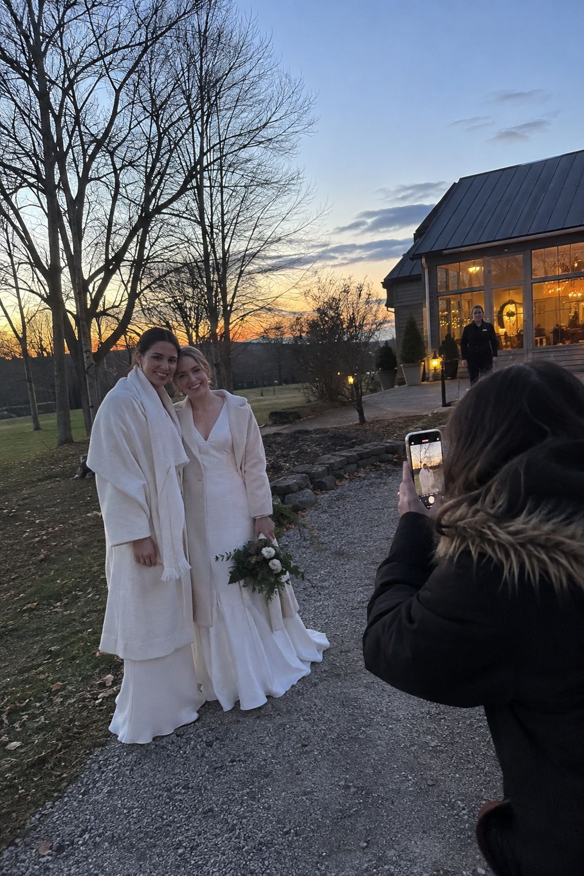 Two brides pause for a friend’s phone portrait in the last winter daylight outside their reception.