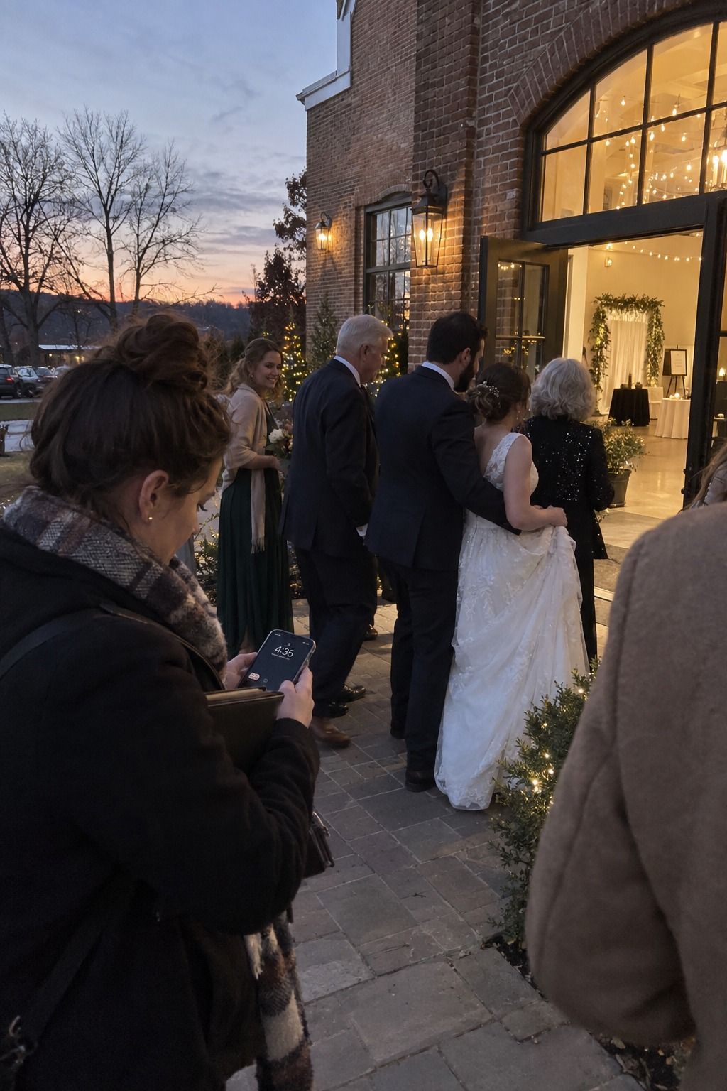 A November wedding coordinator wraps the final outdoor family portrait as daylight fades beside the lit reception entrance.