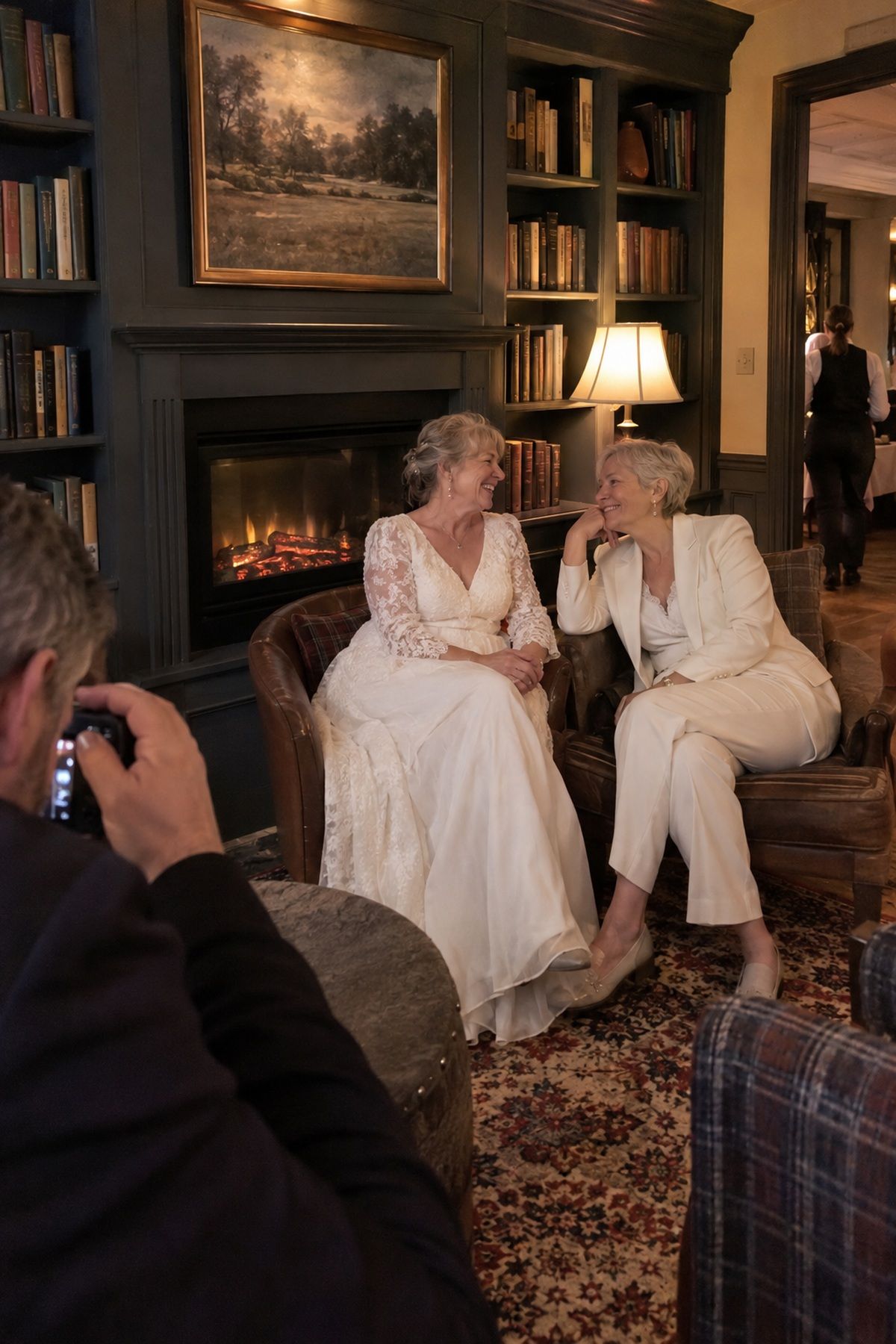Two older brides pause for a portrait beside a sealed fireplace between courses.