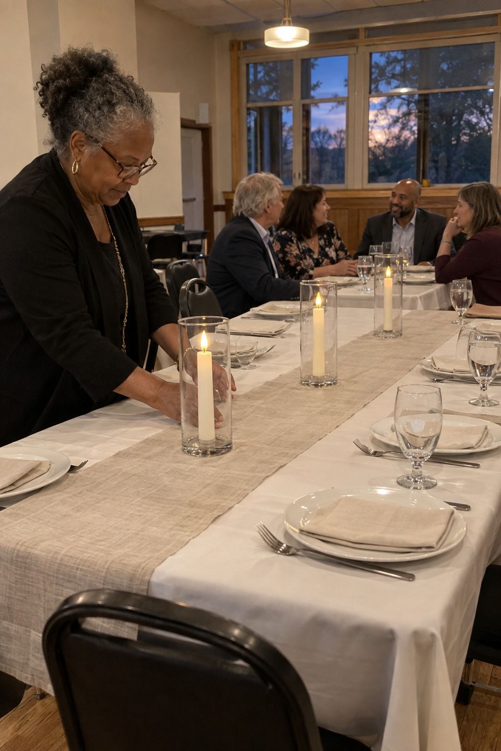 An older Black coordinator checks three stable clear hurricane sleeves around ivory tapers while four guests converse at the far end of a modest hall table.