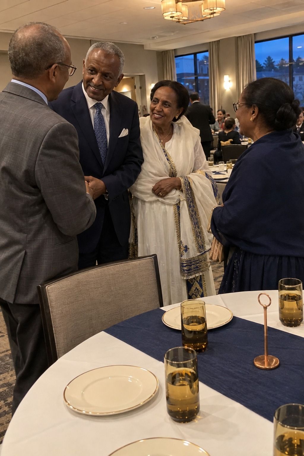 Older East African newlyweds greet friends beside a table whose clearly navy runner softens ivory plates, amber glasses, and one copper detail.