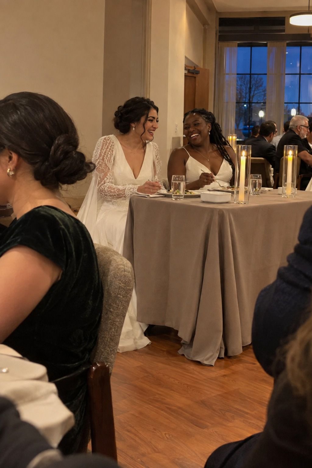 Two brides laugh over dinner at a restrained sweetheart table angled toward guest tables with taupe linen and protected lights.