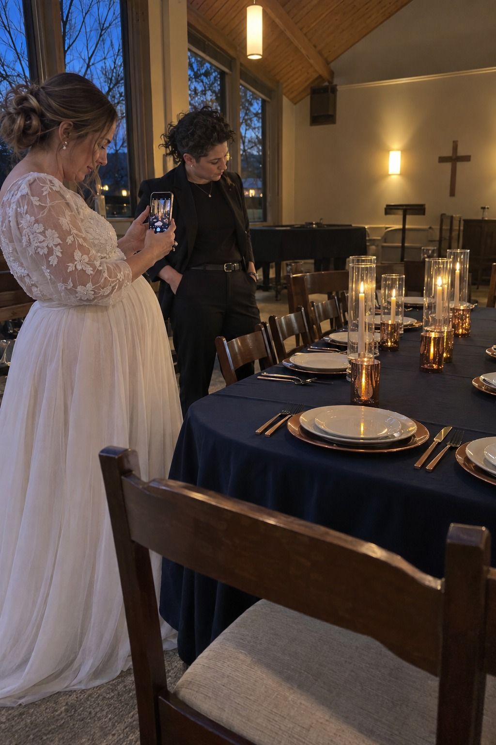 A pregnant bride photographs a completed navy-and-copper table mockup while a nonbinary coordinator inspects it under actual blue-hour and room light.
