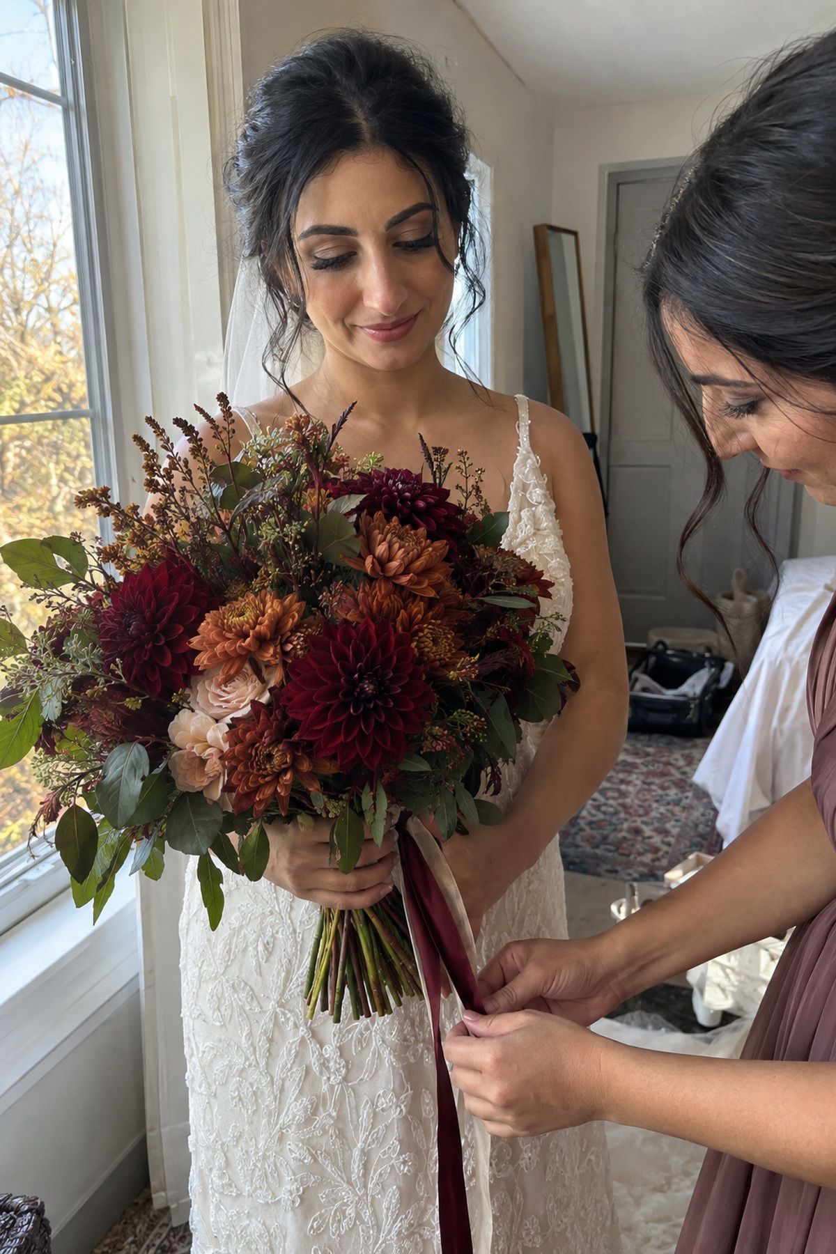 A bride holds a burgundy dahlia and mum bouquet while her sister straightens the ribbon.