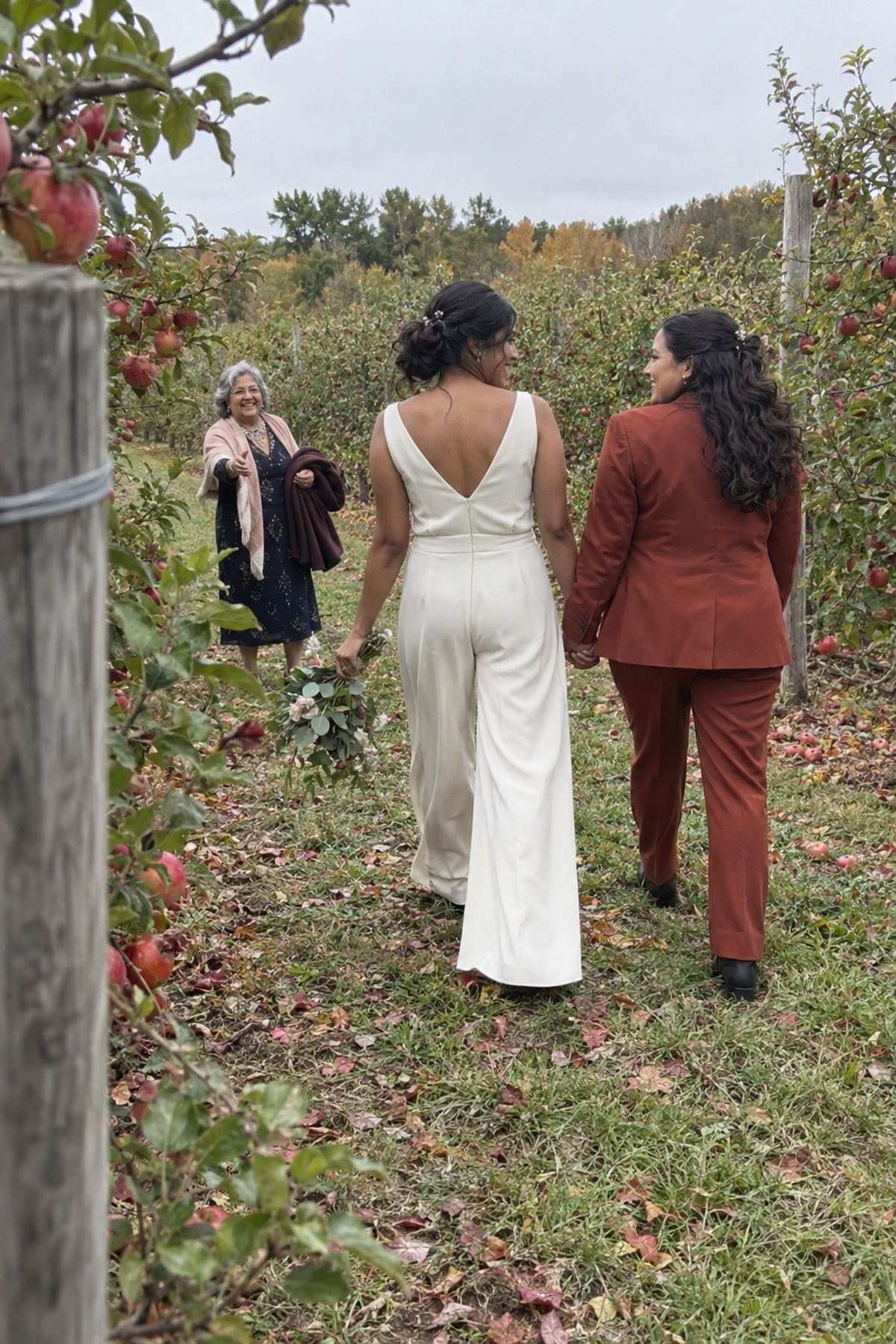Two brides walk between apple rows for October wedding photos.