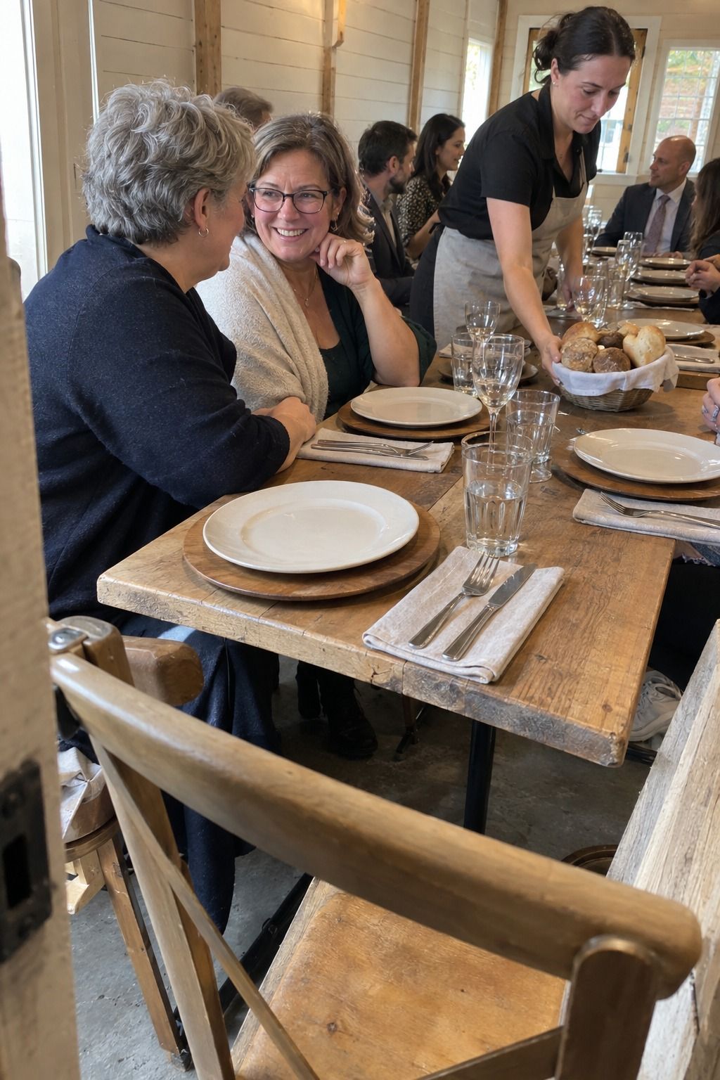 Occupied bright barn-annex table with one natural wooden charger beneath each simple ivory plate.