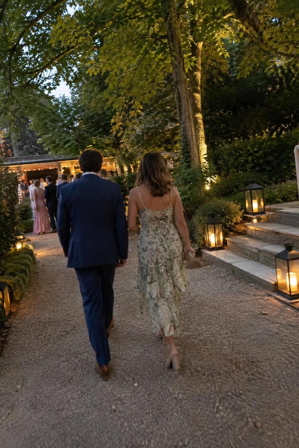 Guests follow closed warm lanterns along a shaded garden path.