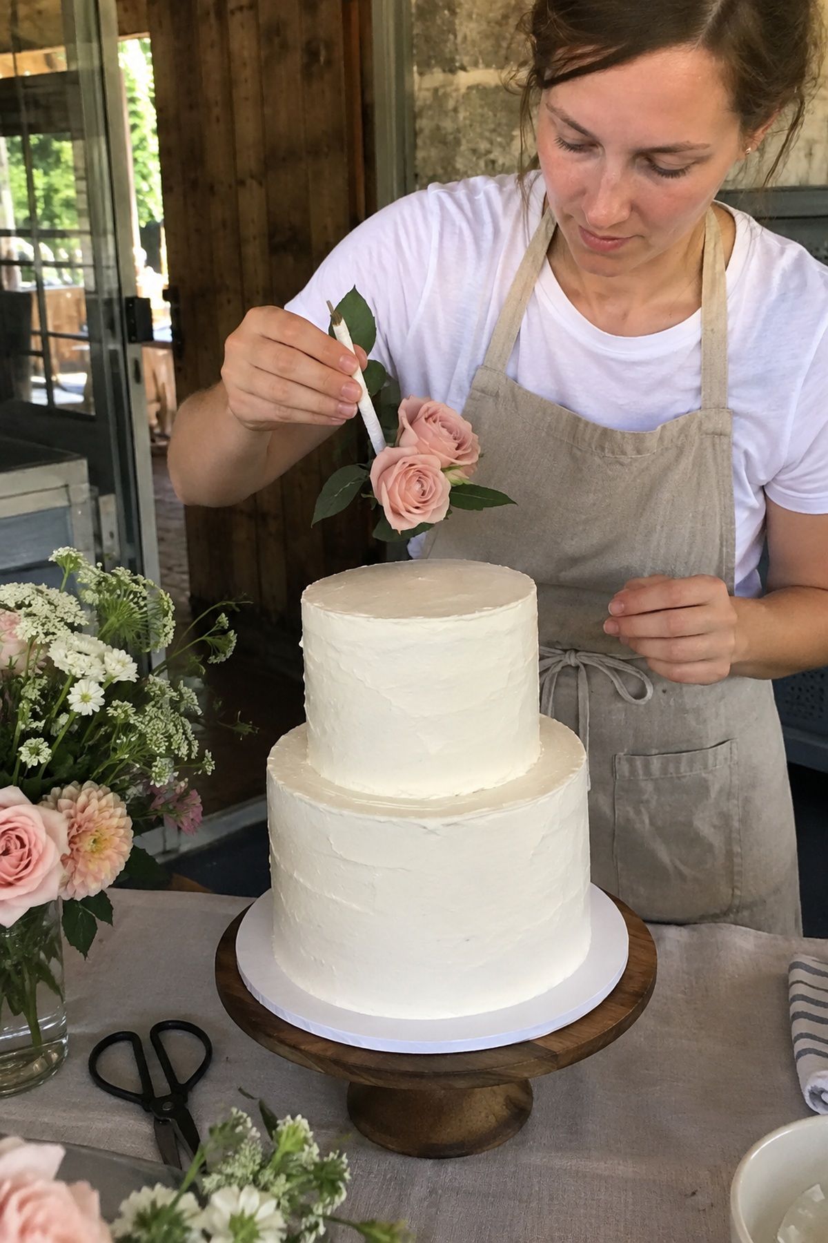 A baker positions fresh blush roses above a shaded summer wedding cake.