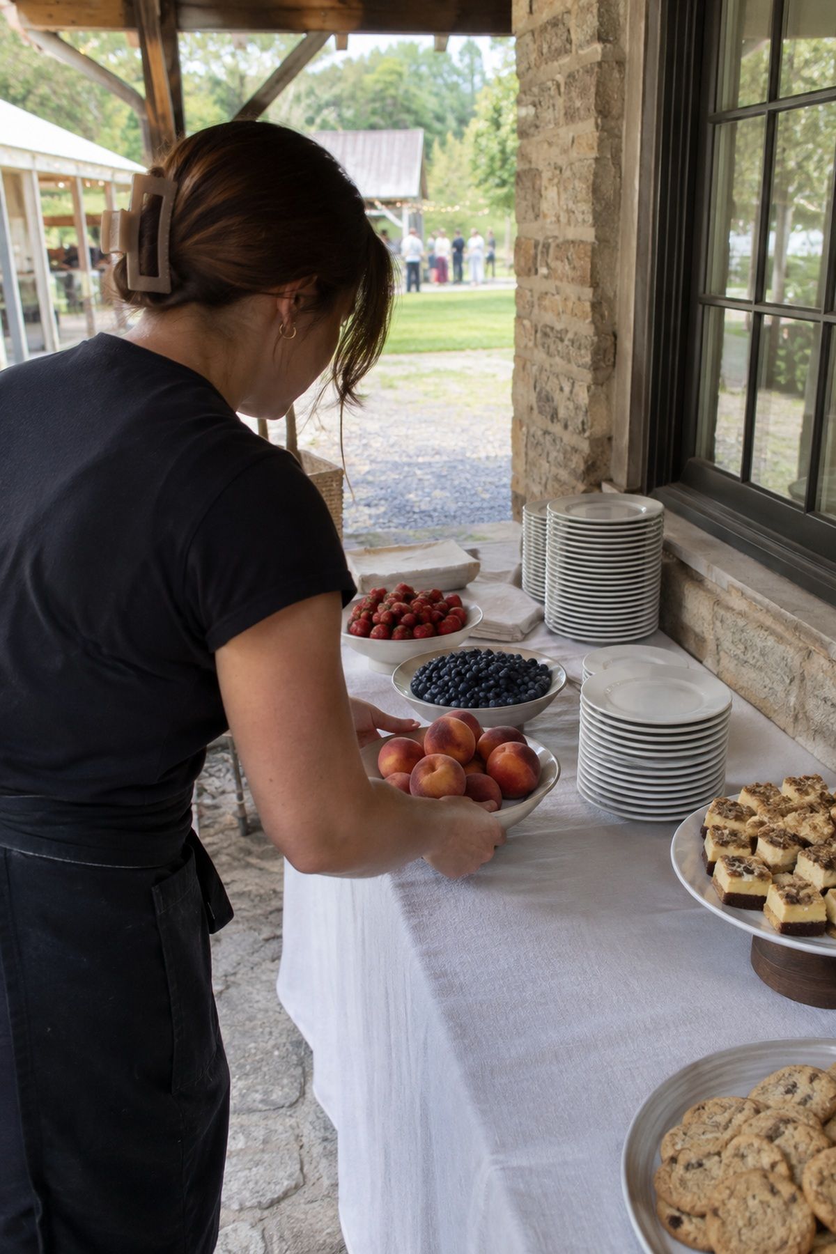 A catering helper adds fresh peaches and summer berries to a shaded wedding dessert table.