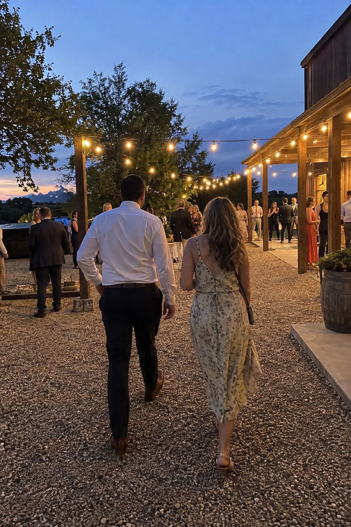 Wedding guests walk safely beneath warm string lights outside a rustic barn at blue hour.