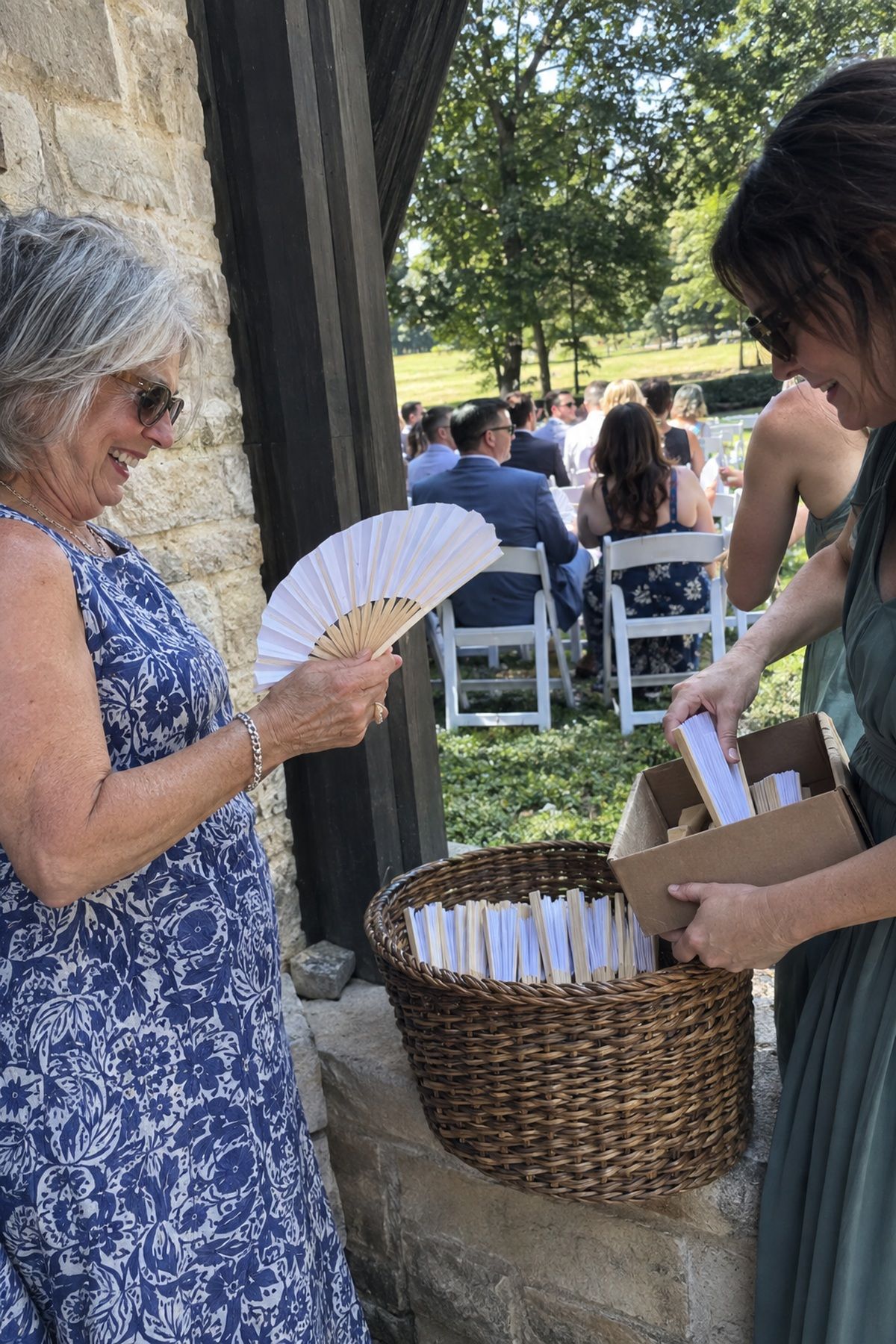 An older wedding guest opens a paper fan from a wicker basket near shaded ceremony seating.