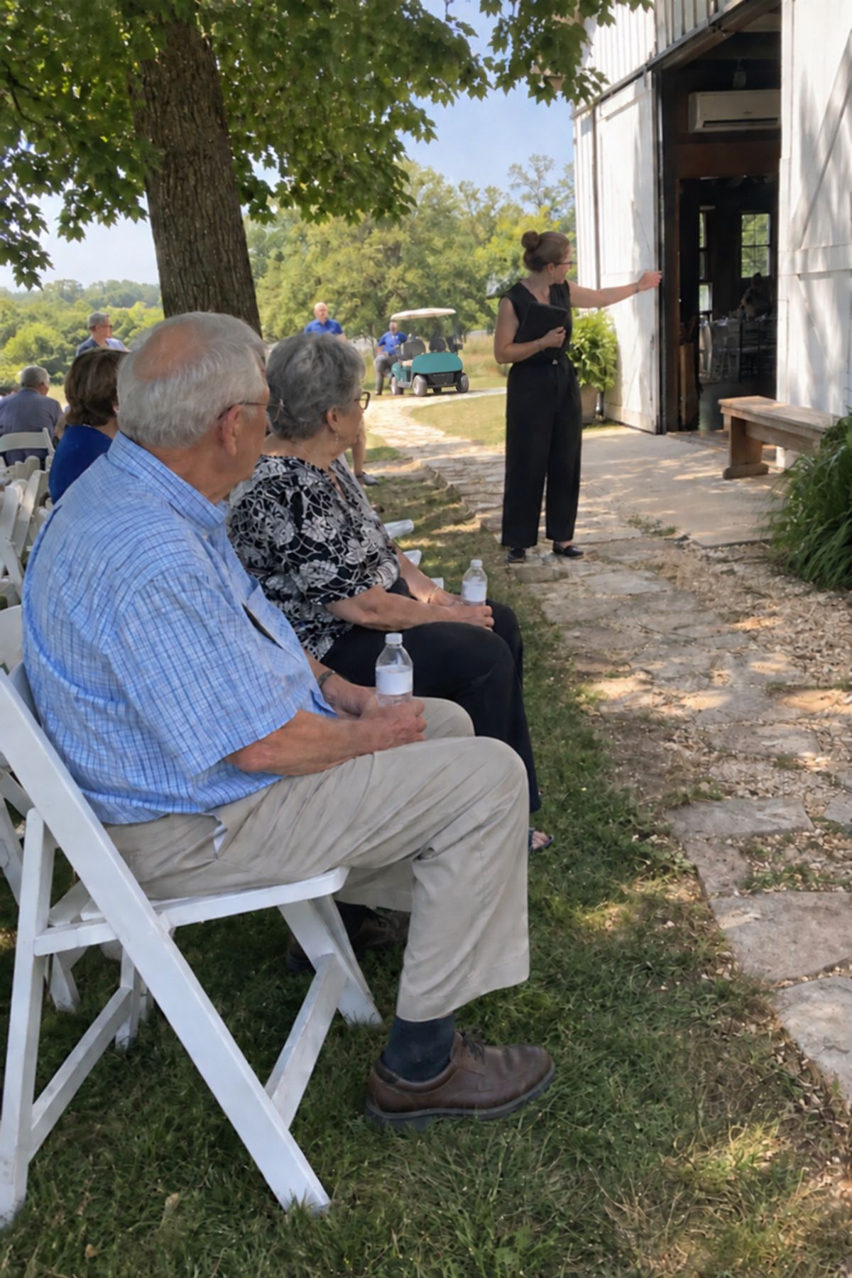 Older wedding guests sit in shade close to an open air-conditioned barn doorway.