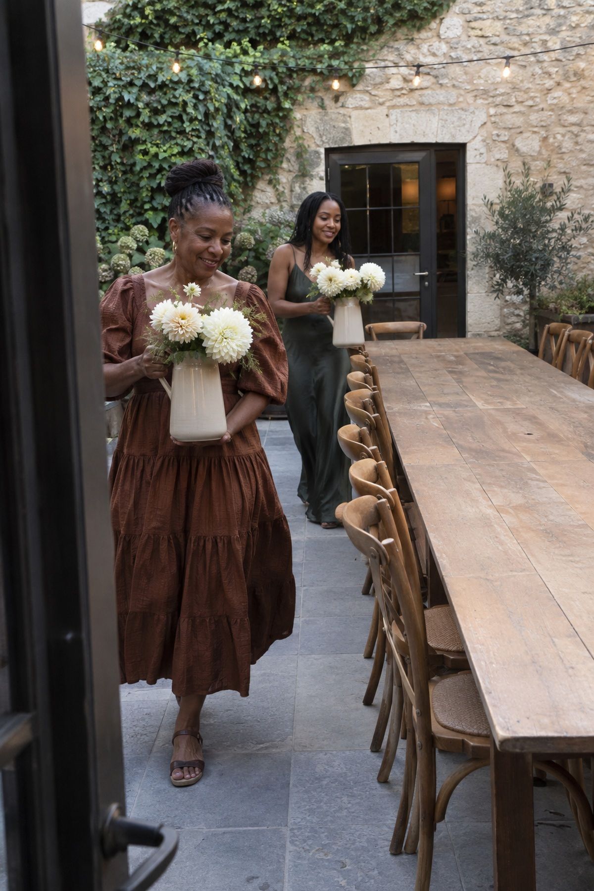 Two wedding helpers carry cream dahlias arranged in stoneware pitchers.