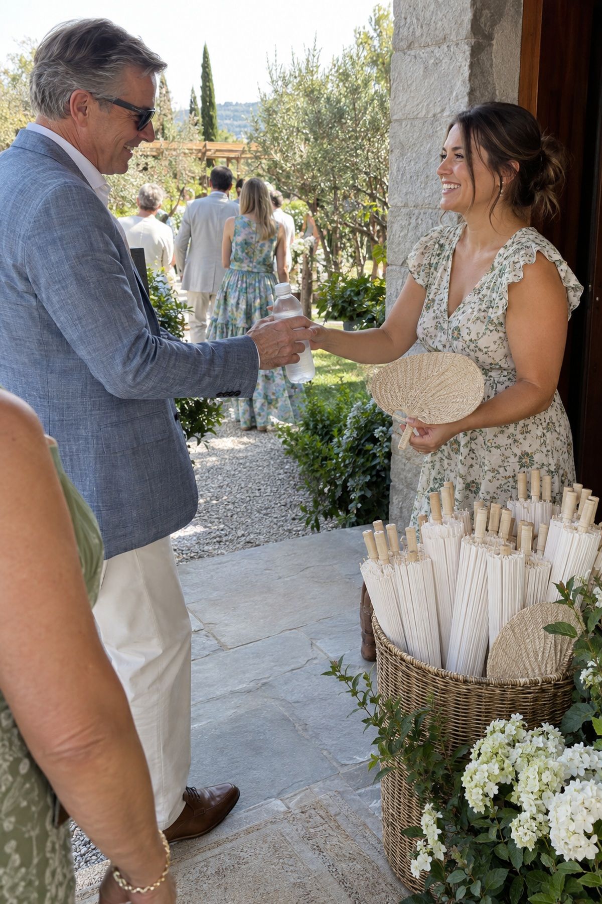 A wedding guest receives a fan and chilled water beside a parasol basket.