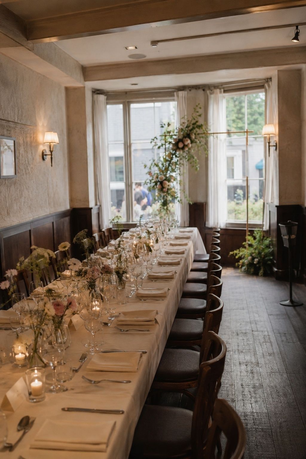 Long restaurant private dining table with white linen, bud vases and candles under tall windows and wall sconces