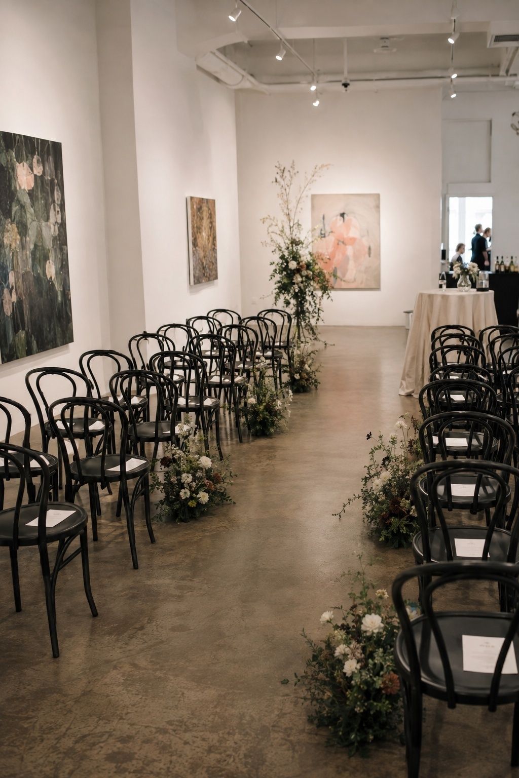 Art gallery wedding ceremony with black bentwood chairs in rows, low white flower arrangements along the aisle, large paintings on white walls and a polished concrete floor