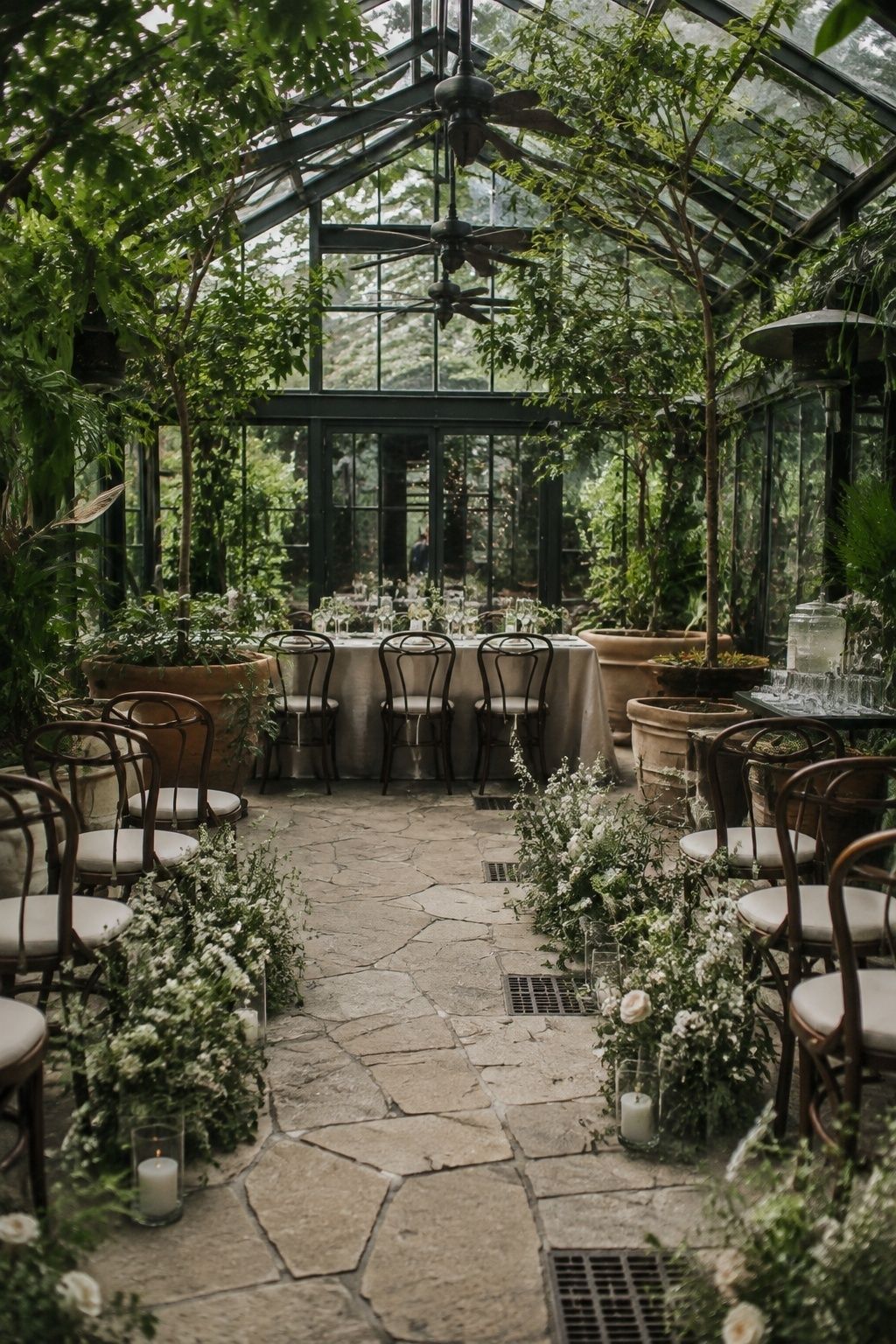 Glass greenhouse wedding with a stone path aisle lined with white flowers, bentwood chairs on both sides and a dining table at the far end among potted plants
