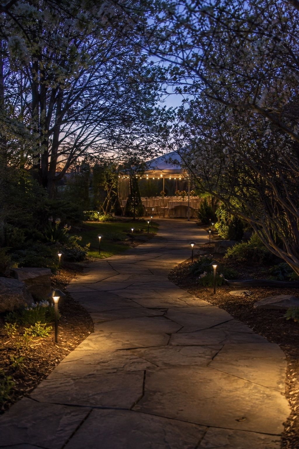 Warm pathway lights along a clear stone route to a garden wedding reception