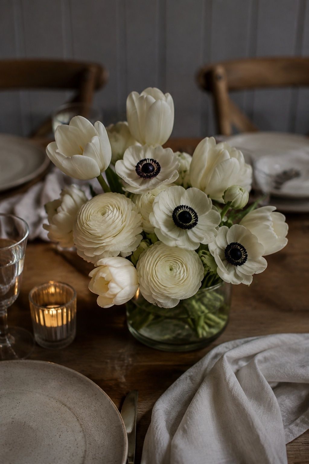 Close diagonal photo of a clear low vessel containing white tulips and pale ranunculus with three black-centered white anemones as graphic accents