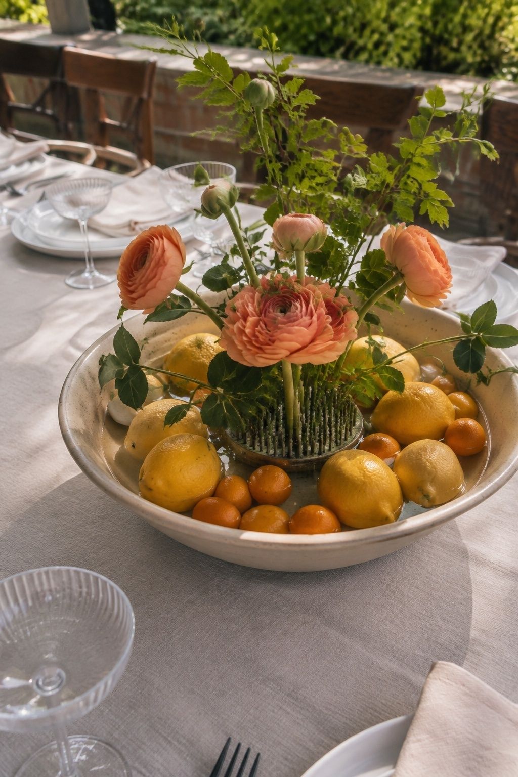 High oblique garden wedding brunch photograph of a shallow bowl with whole lemons and kumquats low around a central water-holding floral insert containing apricot ranunculus and greenery
