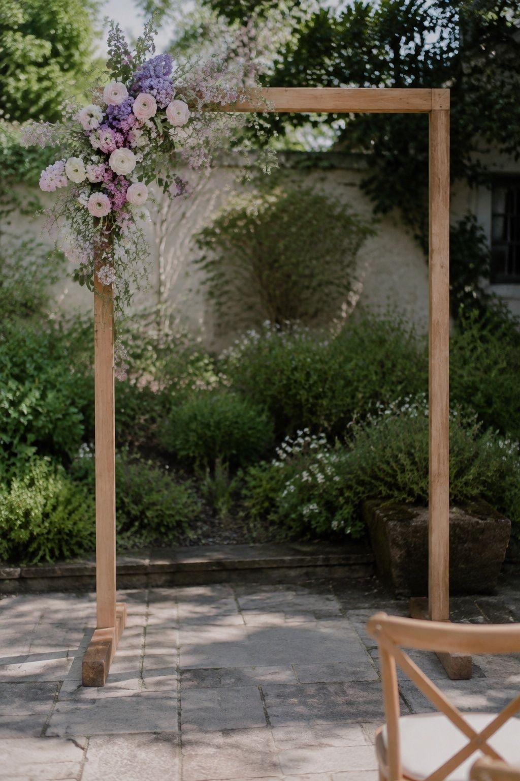 Three-quarter full view of a simple stable wooden arch with a single asymmetrical upper-left corner of lilac, ranunculus and sweet pea, open frame, visible weighted feet on level garden paving