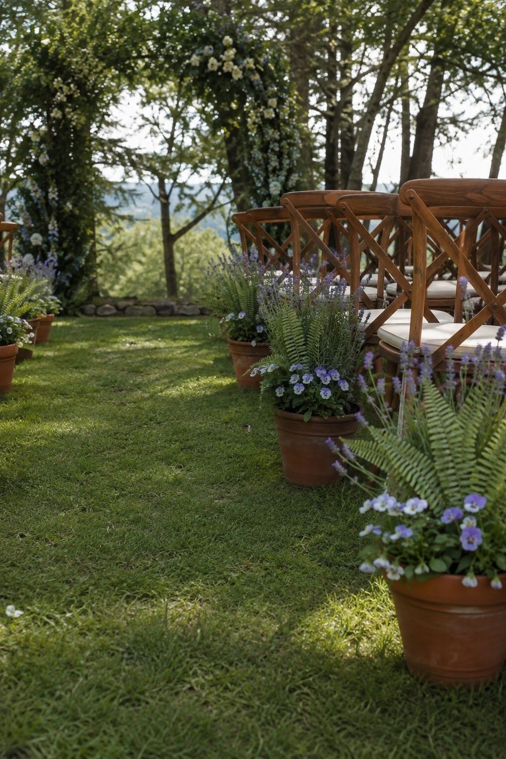 Low oblique view along a broad grass ceremony aisle, heavy terracotta pots of fern, pansies and lavender tucked beside outer chair legs entirely outside walking path