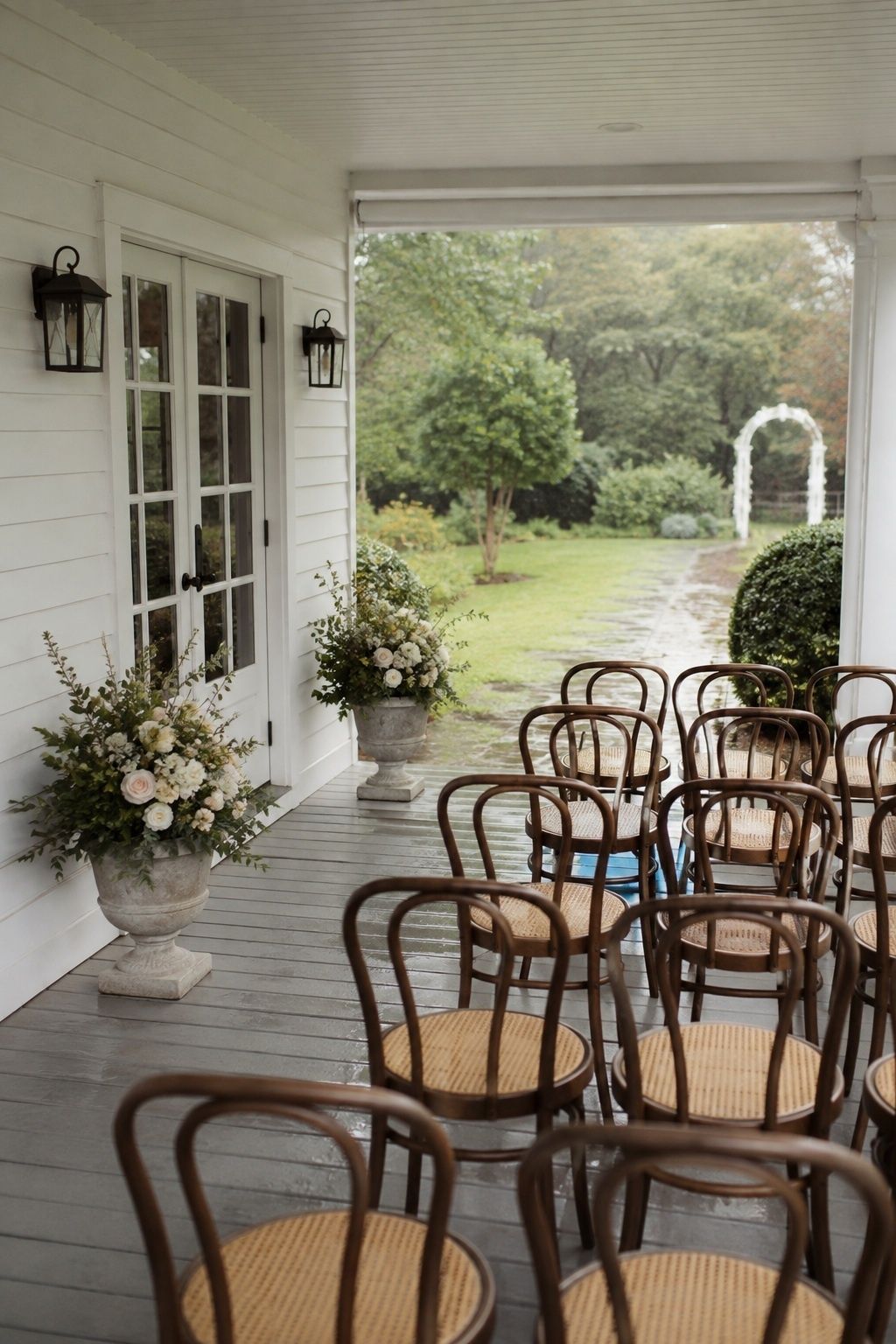 Wide diagonal view of a small covered white farmhouse porch ceremony, two low spring flower urns beside doorway, unobstructed center, three rows of chairs under cover, wet garden beyond