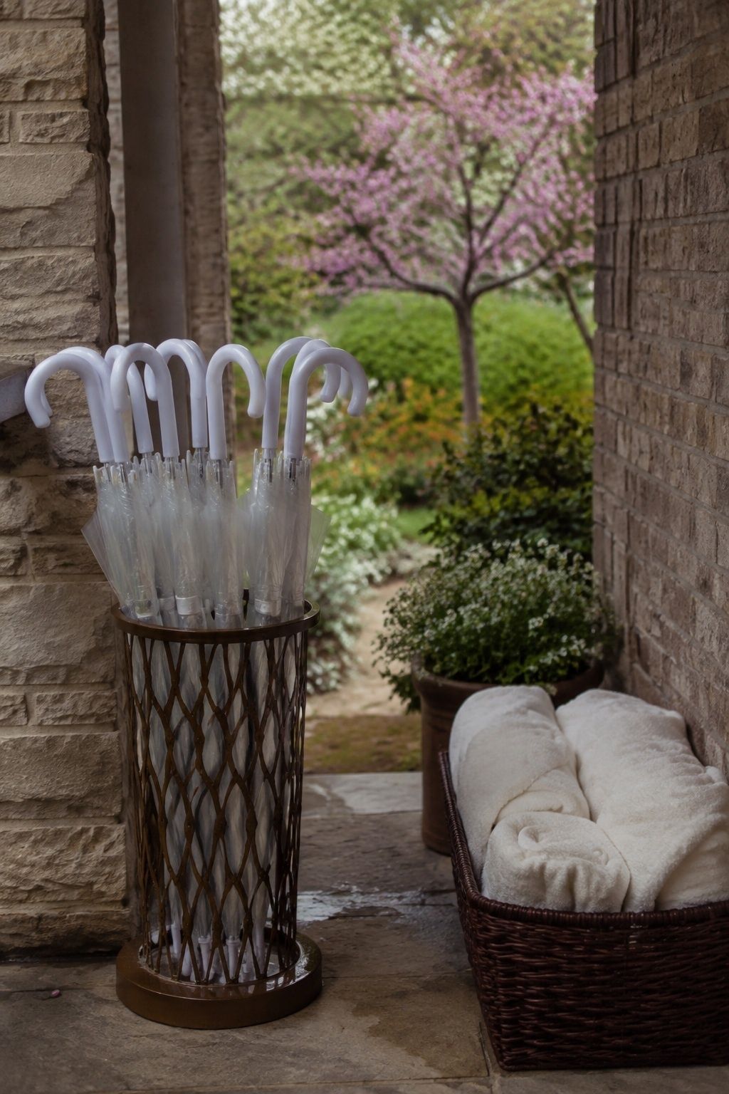 Medium angled view of closed clear dome umbrellas with matching curved handles organized upright in a heavy entrance stand under porch, folded towels in nearby basket, spring garden beyond