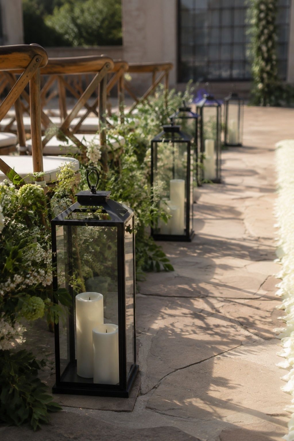 Low diagonal view of black metal glass lanterns containing unlit ivory battery candles, placed outside ceremony walking path beside short chair rows, spring courtyard late afternoon