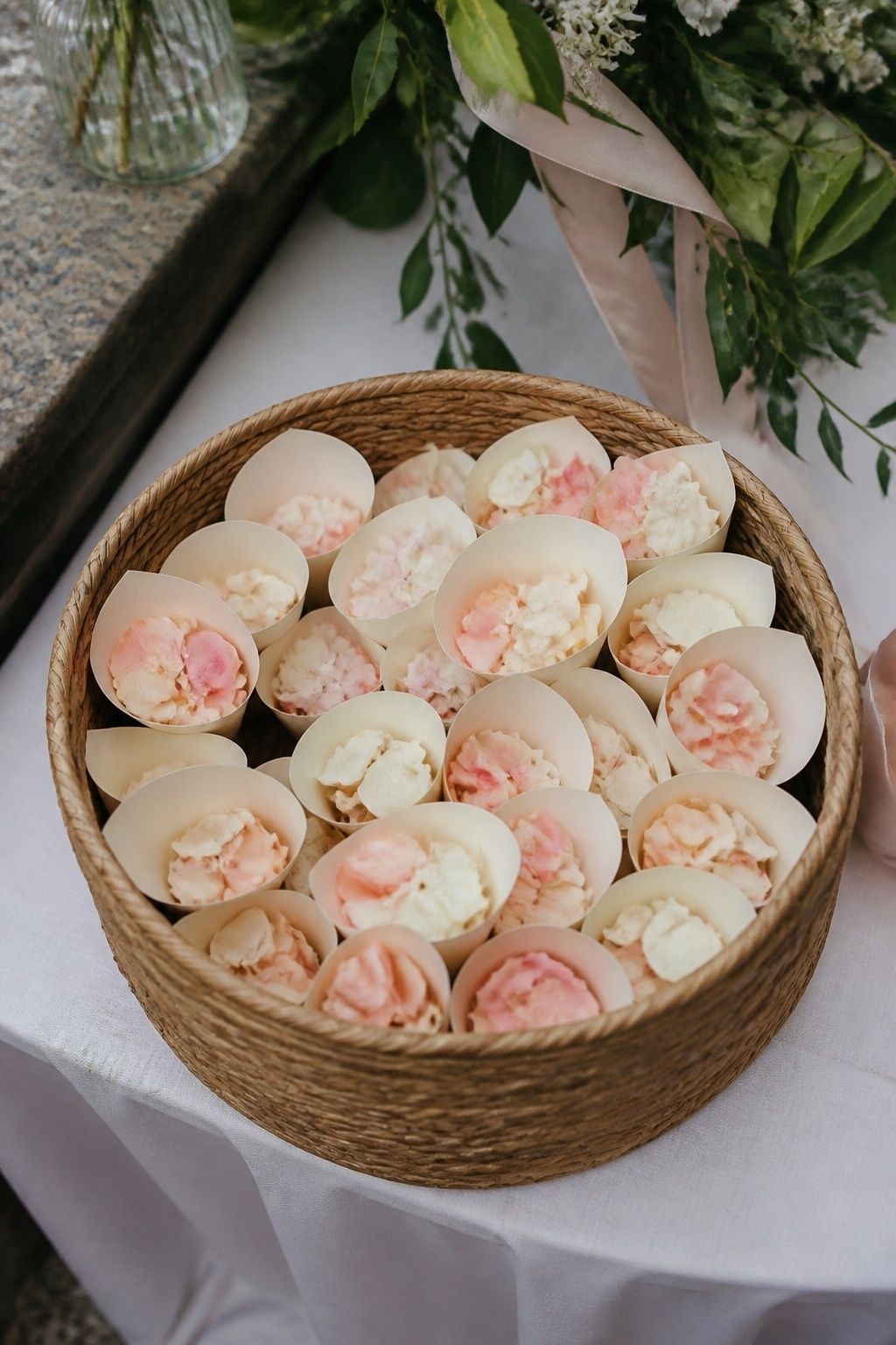 Overhead detail of small cream paper flower cones filled with fresh blush and ivory petals arranged upright in a woven basket on a covered ceremony exit table, ribbon and spring foliage nearby, no text