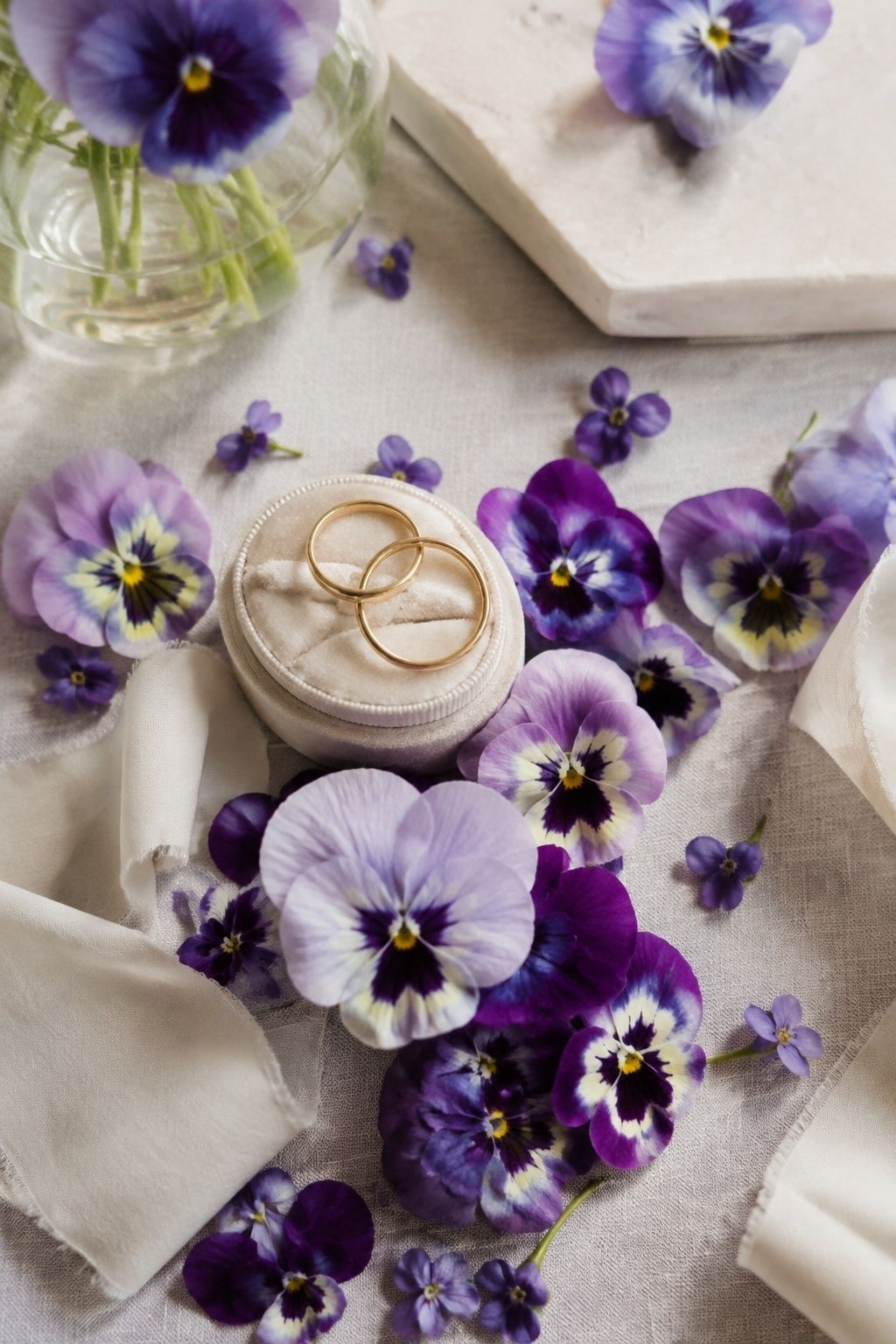 Purple pansies and violets around two gold wedding rings on a cream ring box.
