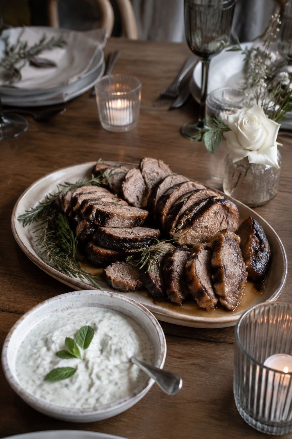 Sliced roasted lamb on a modest shared ceramic platter with rosemary and a separate small mint yogurt bowl