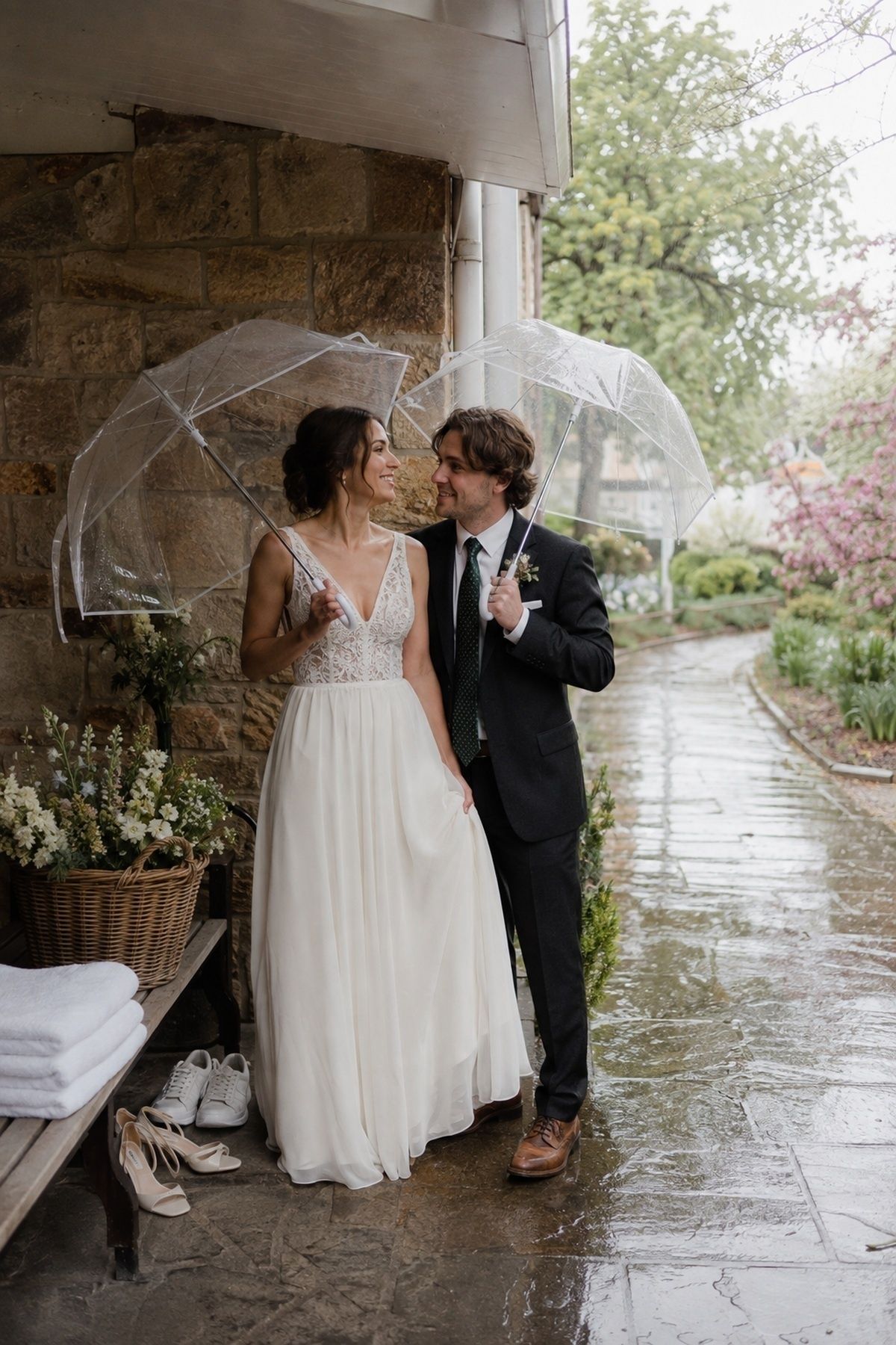 Spring wedding rain plan portrait with clear umbrellas, covered porch, wet stone path, towels, backup shoes, and garden flowers