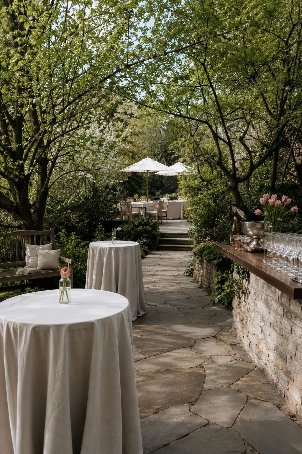 Wide angled garden cocktail setup with two high-top tables, shaded bench, compact bar and stone path to dinner
