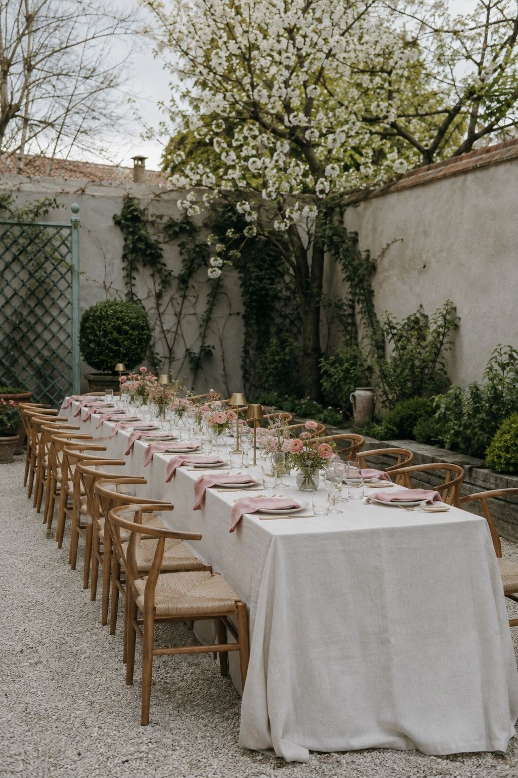 Eye-level diagonal view of one long spring courtyard dinner table for 16 with low flowers, small lamps and clear circulation
