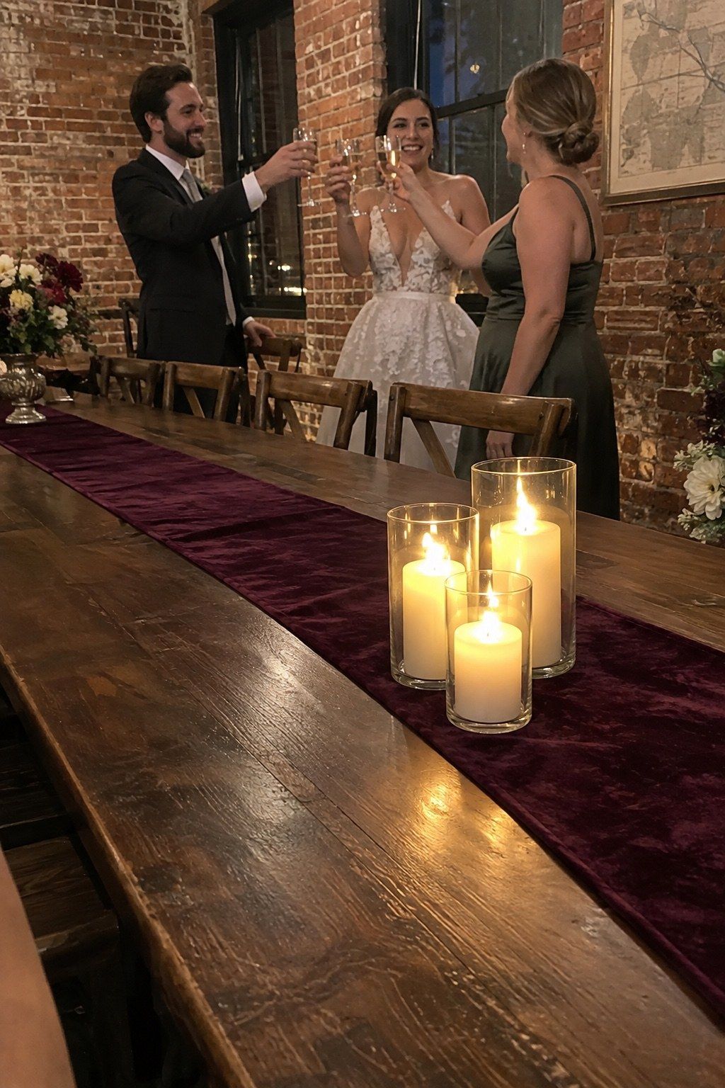 Guests toast beyond a plum velvet runner lit by three stable enclosed glass candles, with florals kept at the table ends.