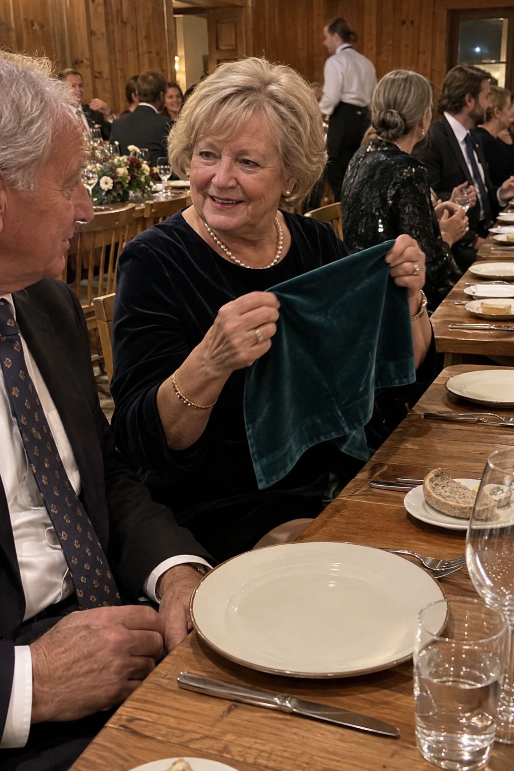 An older guest unfolds a teal velvet napkin at chest height beside a clean empty plate before service.