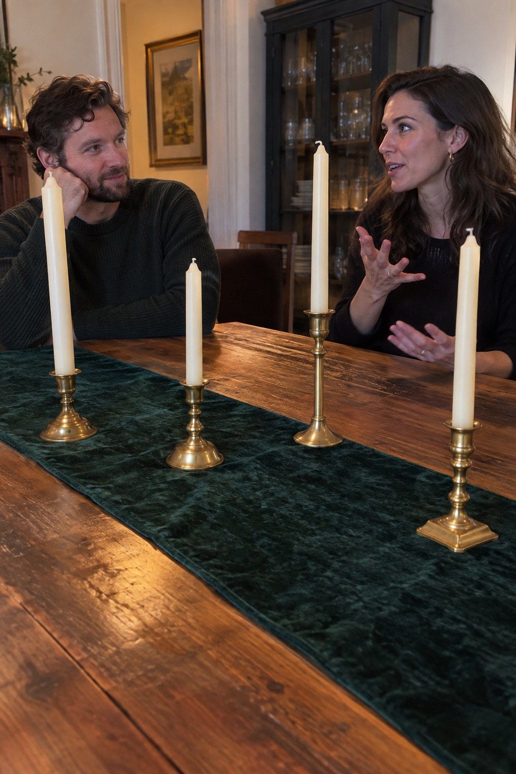 Two guests converse across a deep green runner with safely spaced unlit brass tapers.