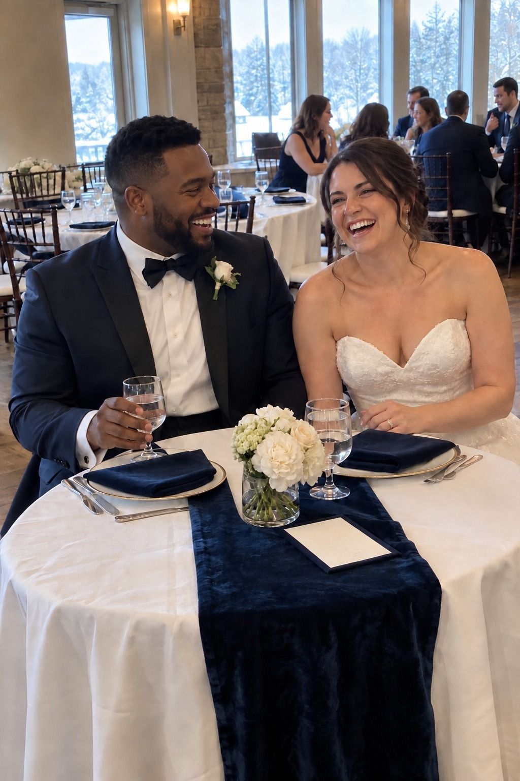A newlywed couple laughs naturally behind a navy velvet sweetheart-table runner and compact white flowers.