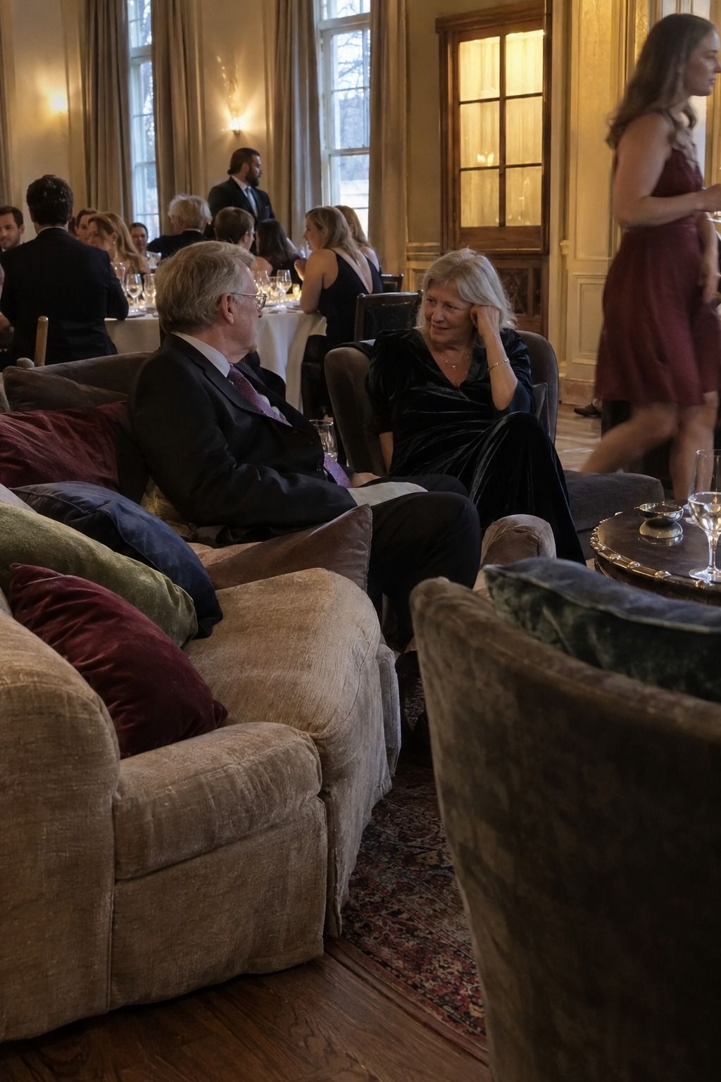 Older guests talk in a genuinely occupied lounge with burgundy, moss and navy velvet pillows beside reception tables.