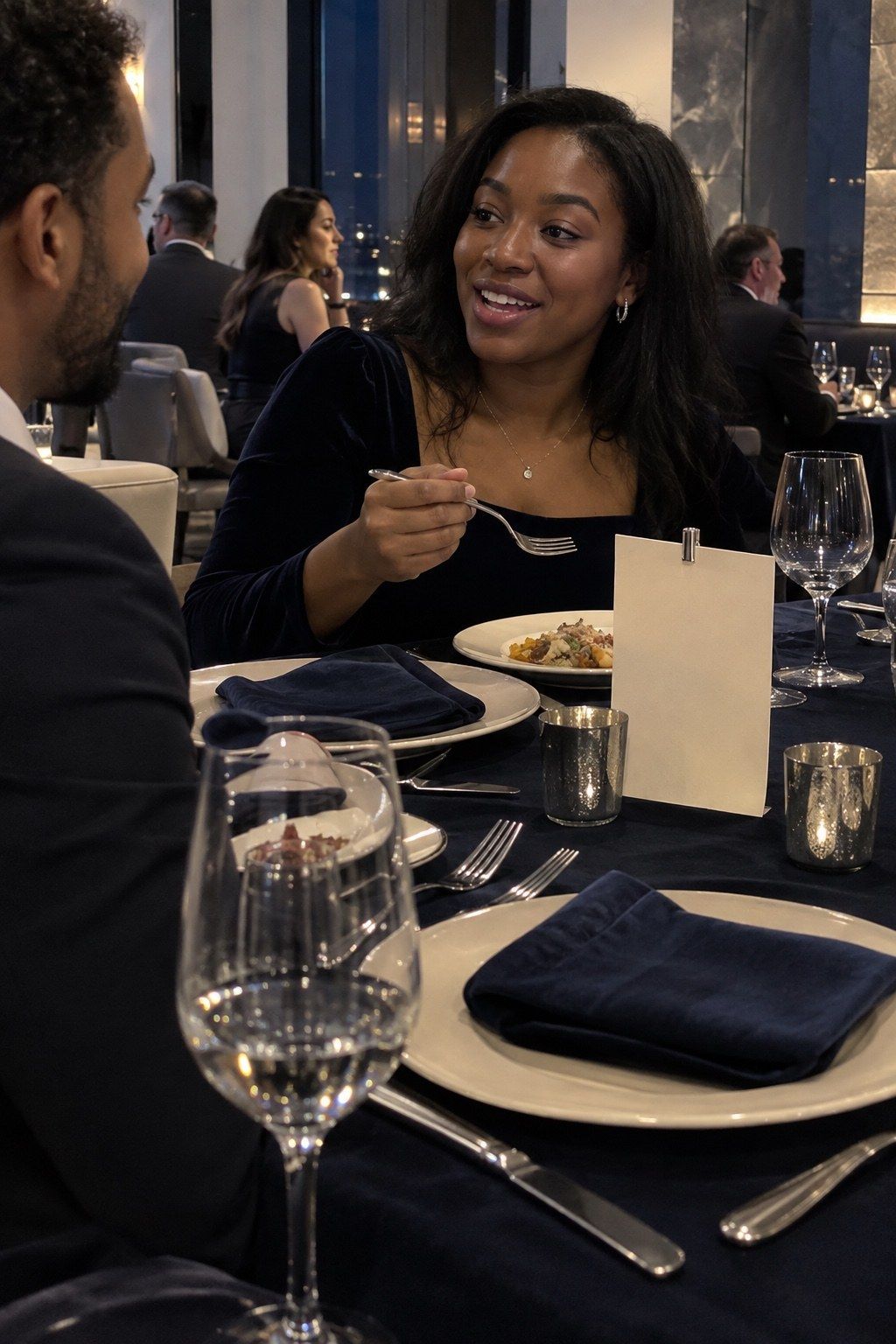 A guest uses silver flatware beside navy velvet, an unprinted clipped card and selective silver vessels.