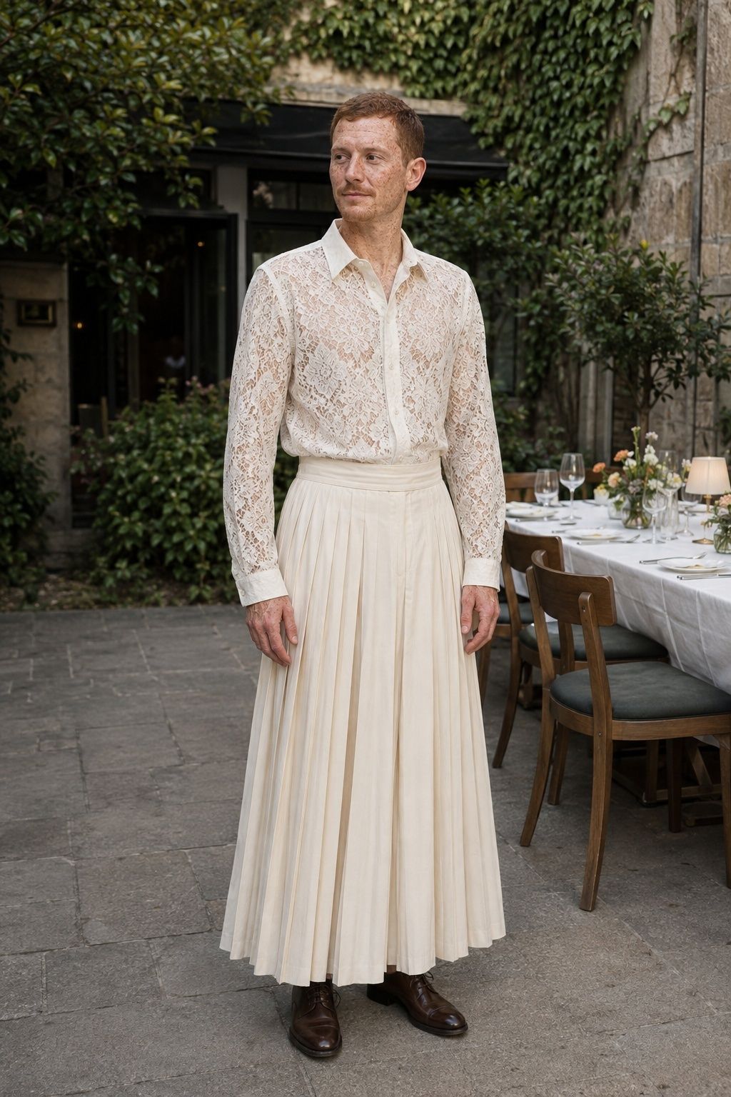 Man in an ivory lace shirt and full pleated skirt at a courtyard reception.